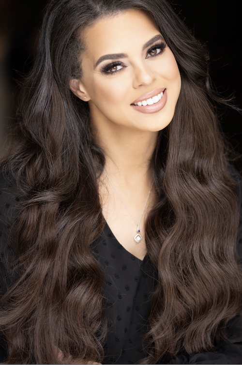 Professional portrait of a woman with long wavy dark hair, soft glam makeup, and a warm confident smile.