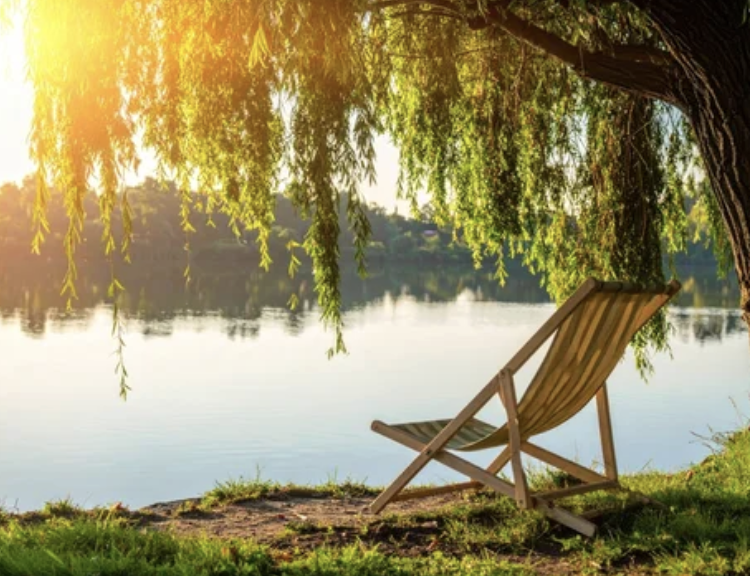 Wooden chair resting beneath a willow tree by a peaceful lake at sunset, symbolizing rest, reflection, and renewal in faith.